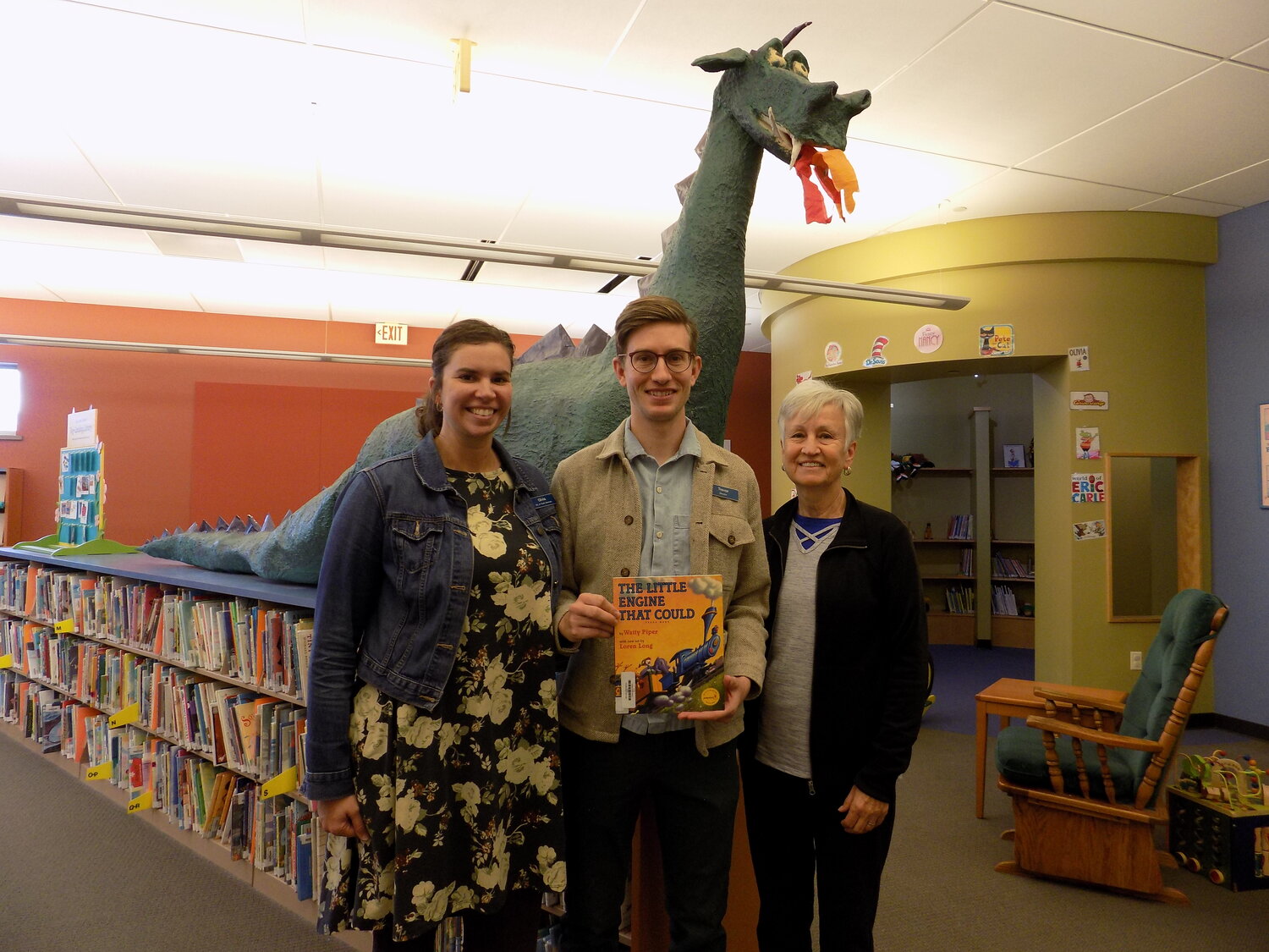 Olivia Kahler (Director of Youth Services), Trevor Sherping (Library Director) and Shirley Harland (KPL Foundation board member) at the Kalona Public Library, holding the first book title children will receive (“The Little Engline That Could”) when they sign up for Dolly Parton’s Imagination Library.